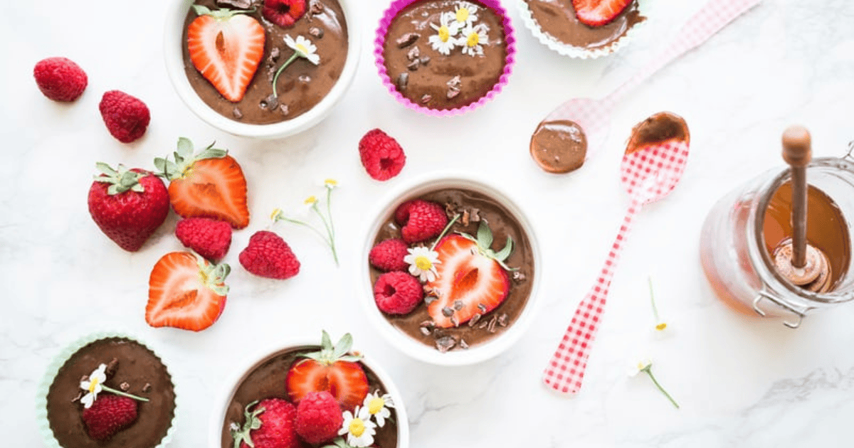 Bowls and cups of chocolate mousse are topped with strawberries, raspberries, cacao nibs, and white flowers. Fresh berries and a jar of chocolate sauce are nearby, with a spoon resting on a checkered napkin—an inviting scene that offers inspiration for those exploring how to reset your relationship with food by savoring mindful moments of indulgence.