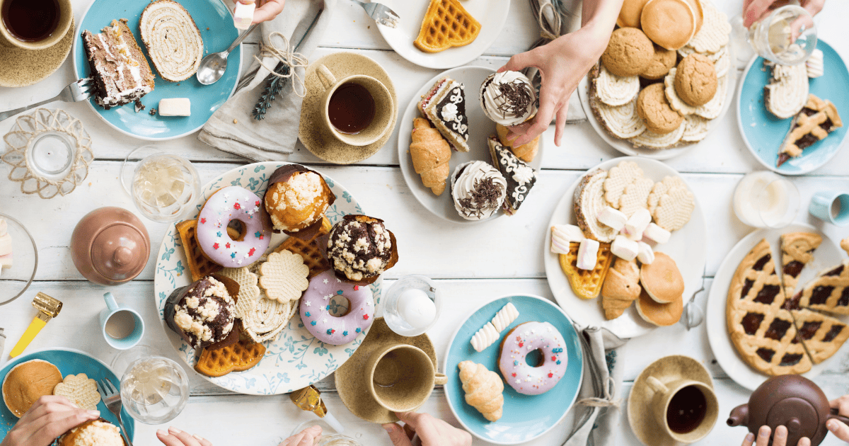 A table filled with assorted desserts, including donuts, ice cream, waffles, cookies, pastries, and cups of coffee and tea—perfect to curb late night cravings—with several hands reaching for treats.