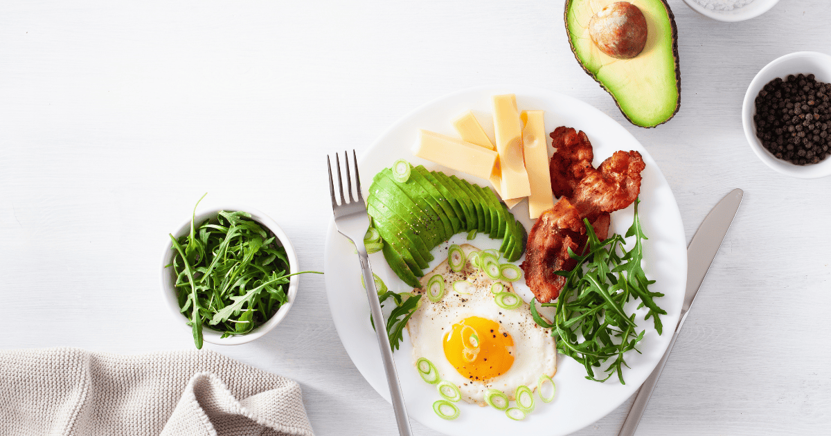 A white plate with a sunny side up egg, sliced avocado, bacon, cheese slices, and arugula—a tempting spread for any breakfast lover. Nearby are a bowl of arugula, half an avocado, black pepper, and a fork and knife. It’s the kind of meal that makes you wonder: has keto ruined my relationship with food or inspired me to see healthy eating in a whole new.