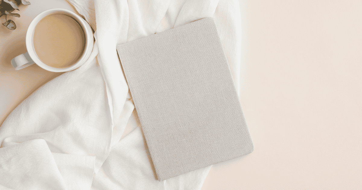A beige notebook rests on a soft white fabric next to a cup of coffee with milk, creating a calm, cozy scene on a light-colored surface—perfect for jotting down affirmations for healthy eating.