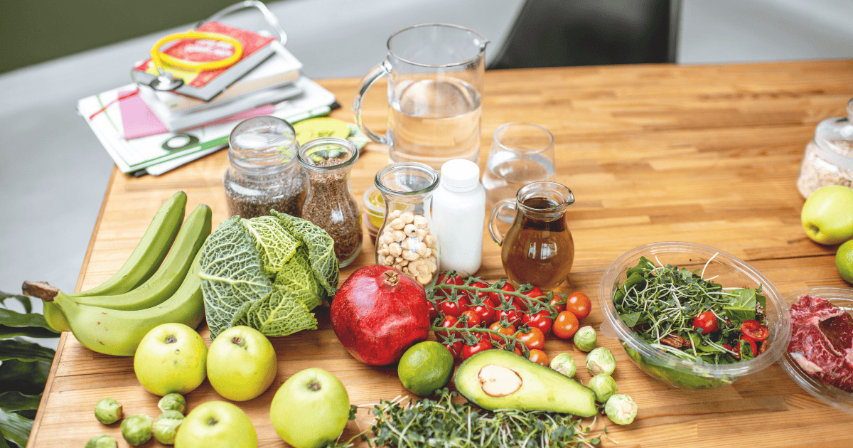 A wooden table with various fruits, vegetables, nuts, seeds, glass jugs of water, a bowl of salad, books, and folders—creating an inviting scene that might be found in the workspace of someone exploring healthy lifestyles. Visible items include bananas, apples, avocado, cabbage, cherry tomatoes, and a pomegranate—perfect props for discussing topics like Dietitian vs nutritionist and their approaches to.