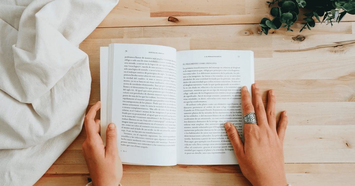 A person with a silver ring on one finger reads an open book on intuitive eating books at a light wooden table, with a white cloth and green plant nearby.