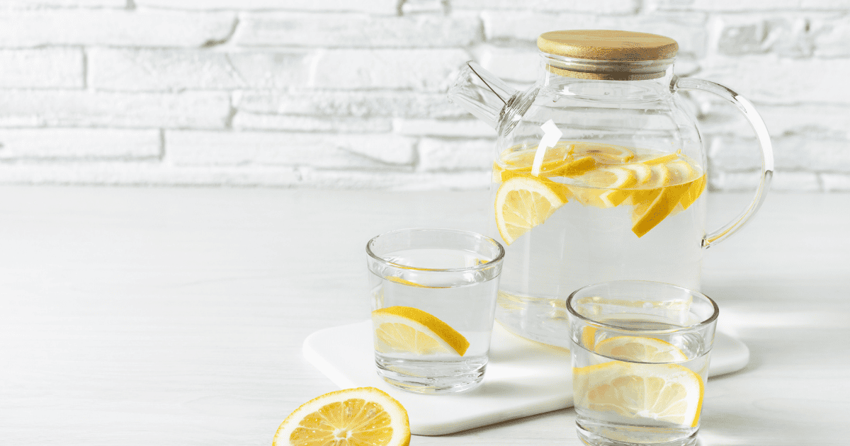 A glass pitcher and two glasses filled with water and lemon slices sit on a white surface with a white brick wall in the background. Another lemon slice lies on the table, offering a refreshing reminder of simple tips to stay hydrated throughout the day.