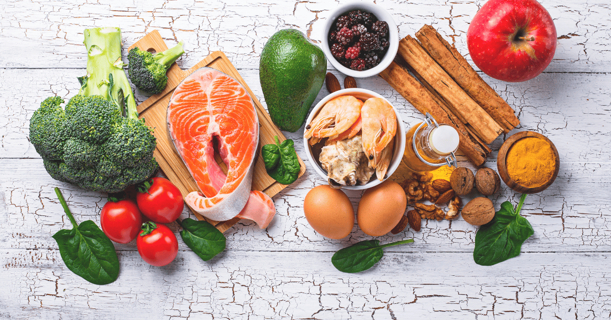 A variety of healthy foods supporting brain health & diet, including salmon, shrimp, eggs, broccoli, tomatoes, spinach, avocado, nuts, blackberries, an apple, cinnamon sticks, and turmeric arranged on a white wooden surface.