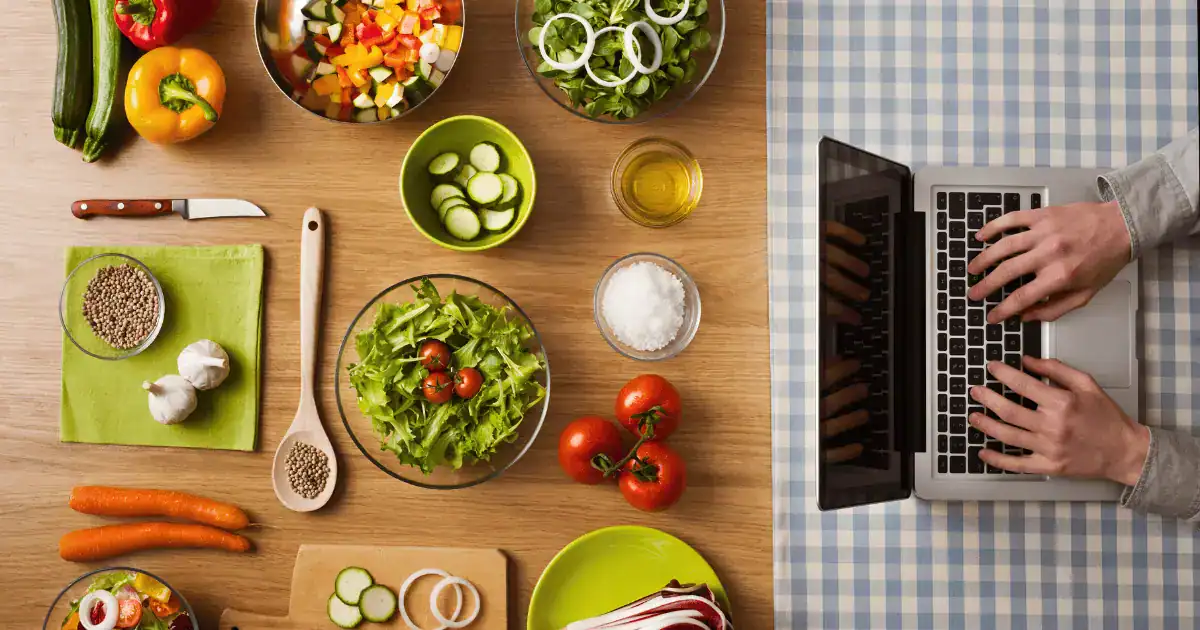 A person types on a laptop next to a table with fresh vegetables, bowls of salad, spices, and cooking ingredients suited for a FODMAP diet on a checkered tablecloth.
