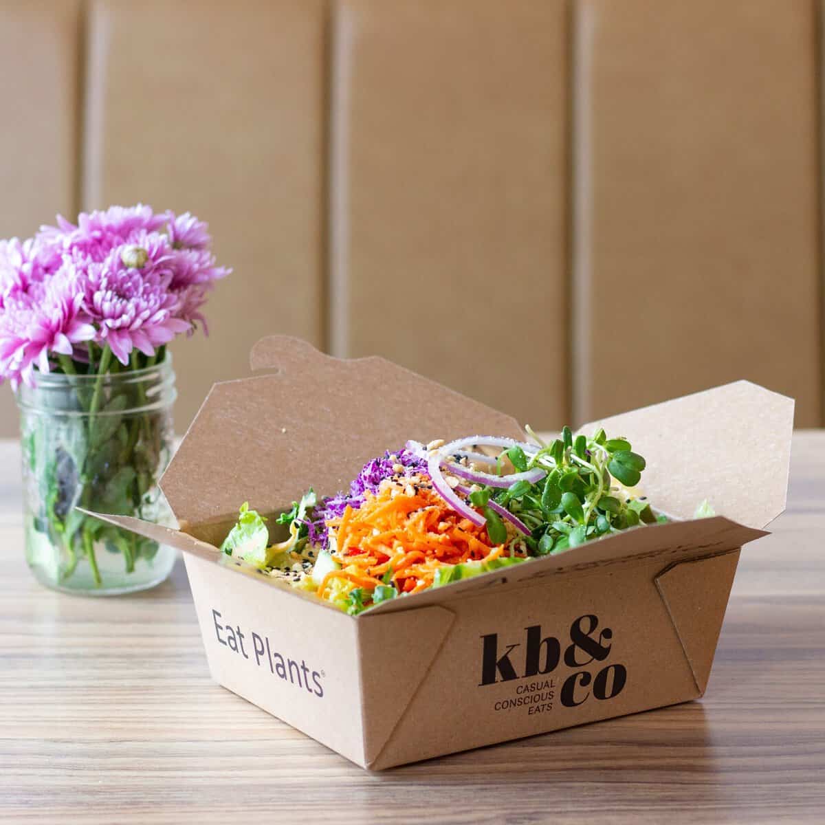 A cardboard takeout box filled with a fresh salad sits on a wooden table next to a glass jar with purple flowers. The box is labeled “kb & co” and “Eat Plants.” Known as one of the top healthy restaurants in Edmonton, kb & co offers vibrant plant-based options perfect for anyone seeking nourishing meals.