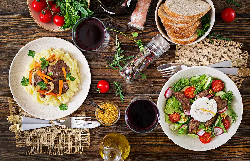 Meal plans by Nutritionist and Dietitian, In good Nutrition. Two plates of food are on a rustic table with bread, wine, tomatoes, condiments, herbs, and utensils.