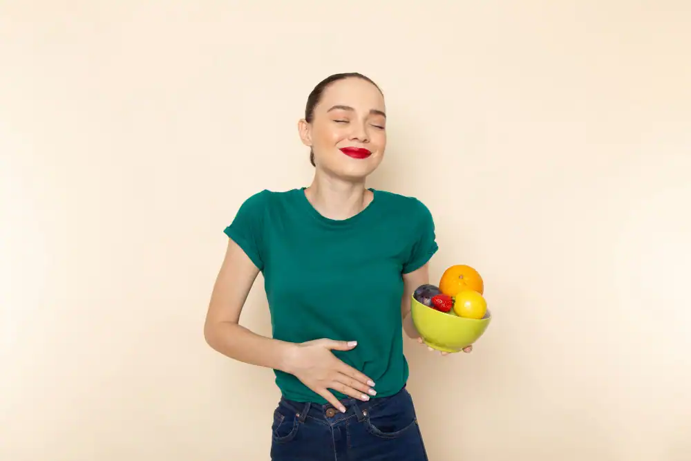 Woman in a green shirt holding a bowl of fruit with one hand and touching her stomach with the other, smiling with eyes closed, standing against a plain light background.