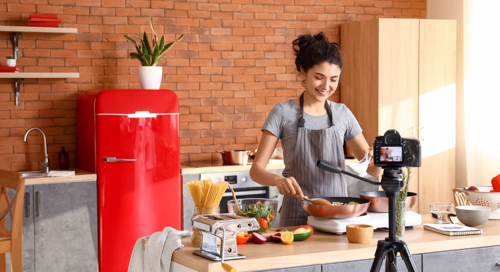 A woman in a gray apron cooks pasta in a kitchen while smiling at a camera on a tripod. The kitchen has a red fridge, potted plant, and ingredients like vegetables and spaghetti on the counter.