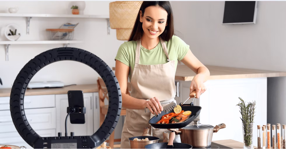 A woman in an apron cooks vegetables in a kitchen while smiling at a camera and ring light, possibly filming a cooking tutorial.