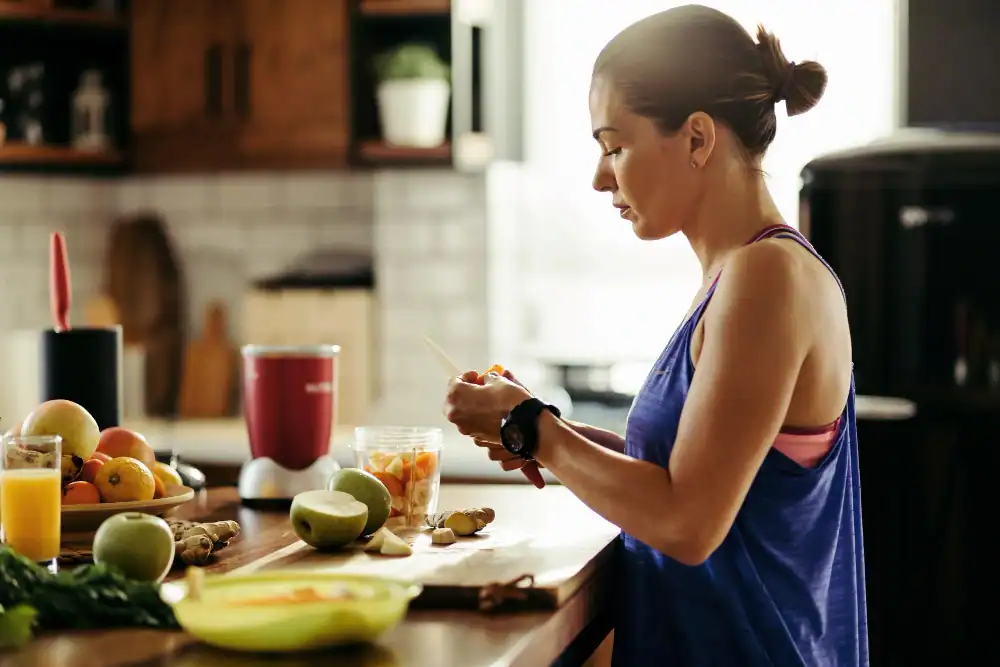 A woman in athletic wear stands in a kitchen, slicing fruit on a cutting board with a blender nearby. Fresh fruits, ginger, and a glass of juice are on the counter. Sunlight streams in from the window.