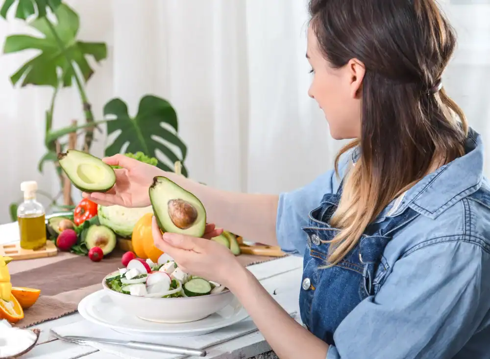 Low FODMAP Dietitian, In Good Nutrition. Woman sitting at a table holds two avocado halves over a salad bowl. Fresh vegetables, a glass bottle, and other ingredients are visible on the table, with plants in the background.