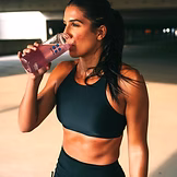 A woman in athletic wear drinks a pink beverage from a clear bottle while standing in an outdoor setting, possibly after exercising.