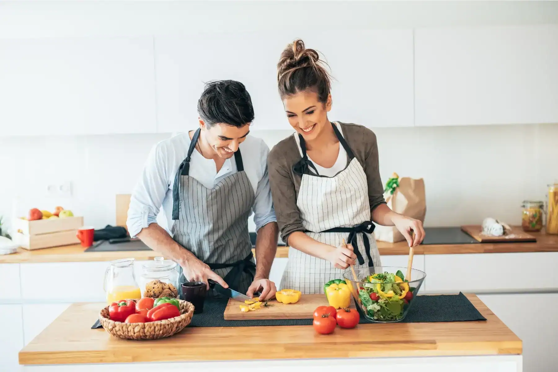 A smiling man and woman wearing aprons prepare food together in a bright kitchen. He slices yellow peppers while she mixes a salad. Fresh vegetables, juice, and cookies are on the counter.