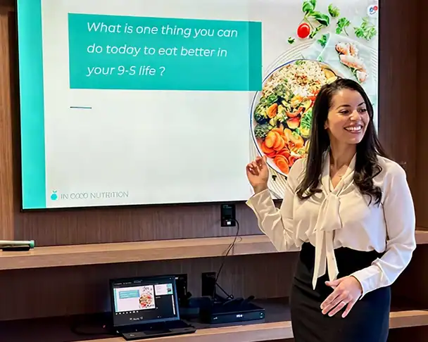 A woman in business attire stands smiling and pointing at a screen displaying the text, “What is one thing you can do today to eat better in your 9-5 life?” with a plate of colorful food pictured.
