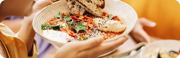 A person holds a bowl of hummus topped with herbs, sesame seeds, chili slices, and pita bread.