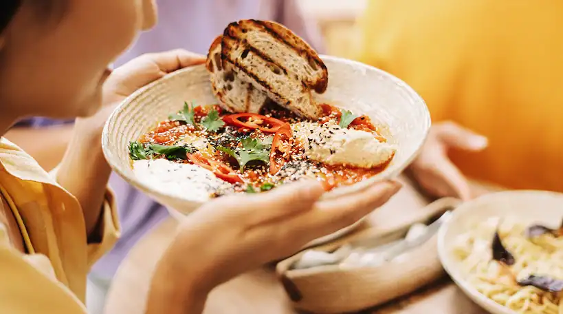 A person holds a bowl of hummus topped with vegetables, sesame seeds, herbs, and sliced chilies, served with two pieces of grilled bread.