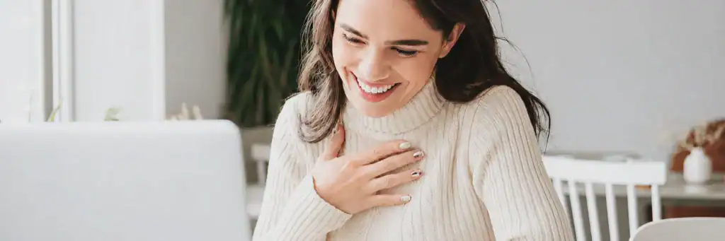 Woman with long brown hair wearing a cream sweater smiles and touches her chest while looking at a laptop, sitting at a desk in a bright indoor space with plants in the background.