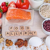 A cutting board with salmon, nuts, milk, eggs, beans, tomatoes, strawberries, and oatmeal, with wooden letter tiles spelling “FOOD ALLERGY.”.