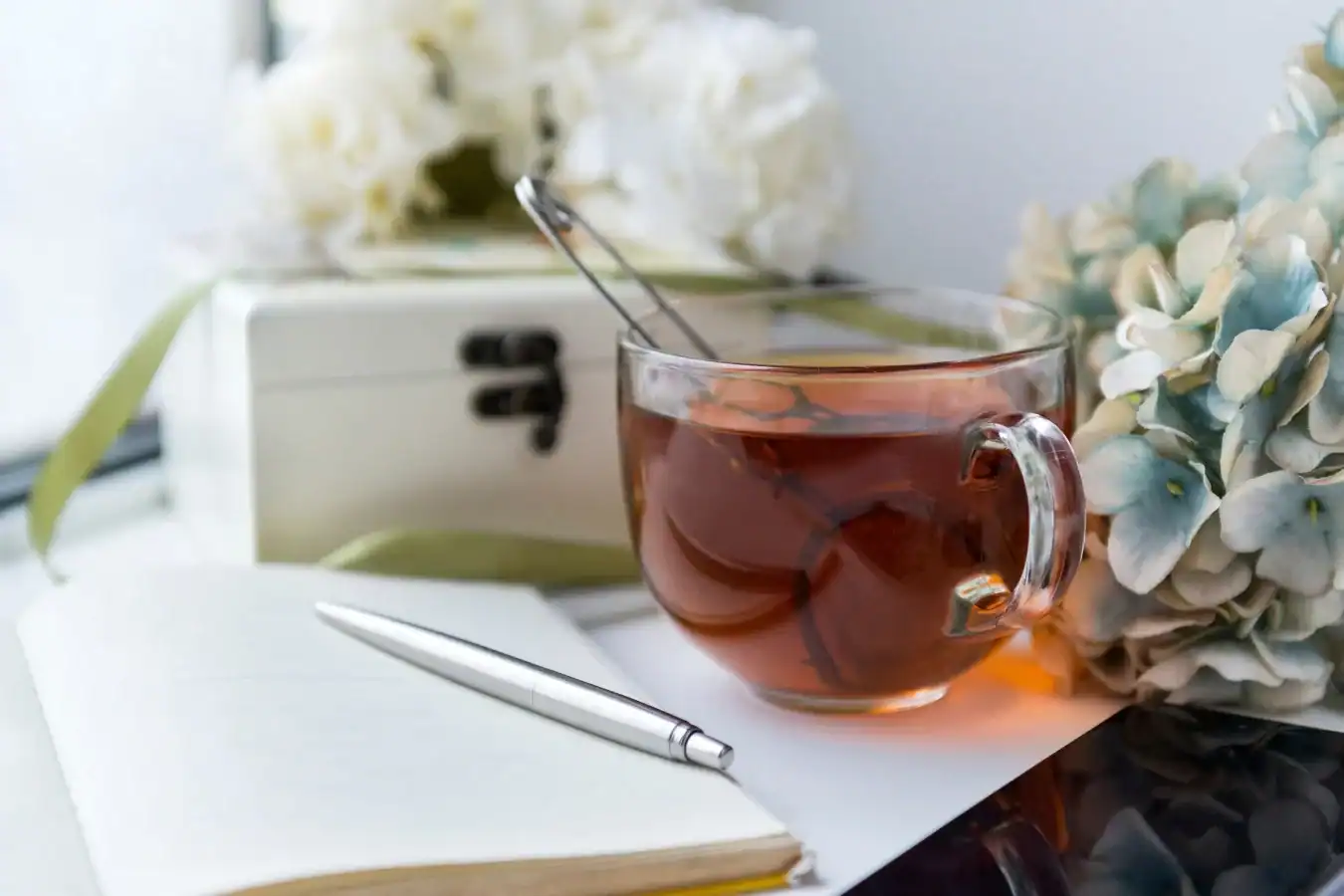 A glass cup of tea with a spoon inside sits beside an open notebook and pen, white flowers, and a decorative box in a calm, softly lit setting.