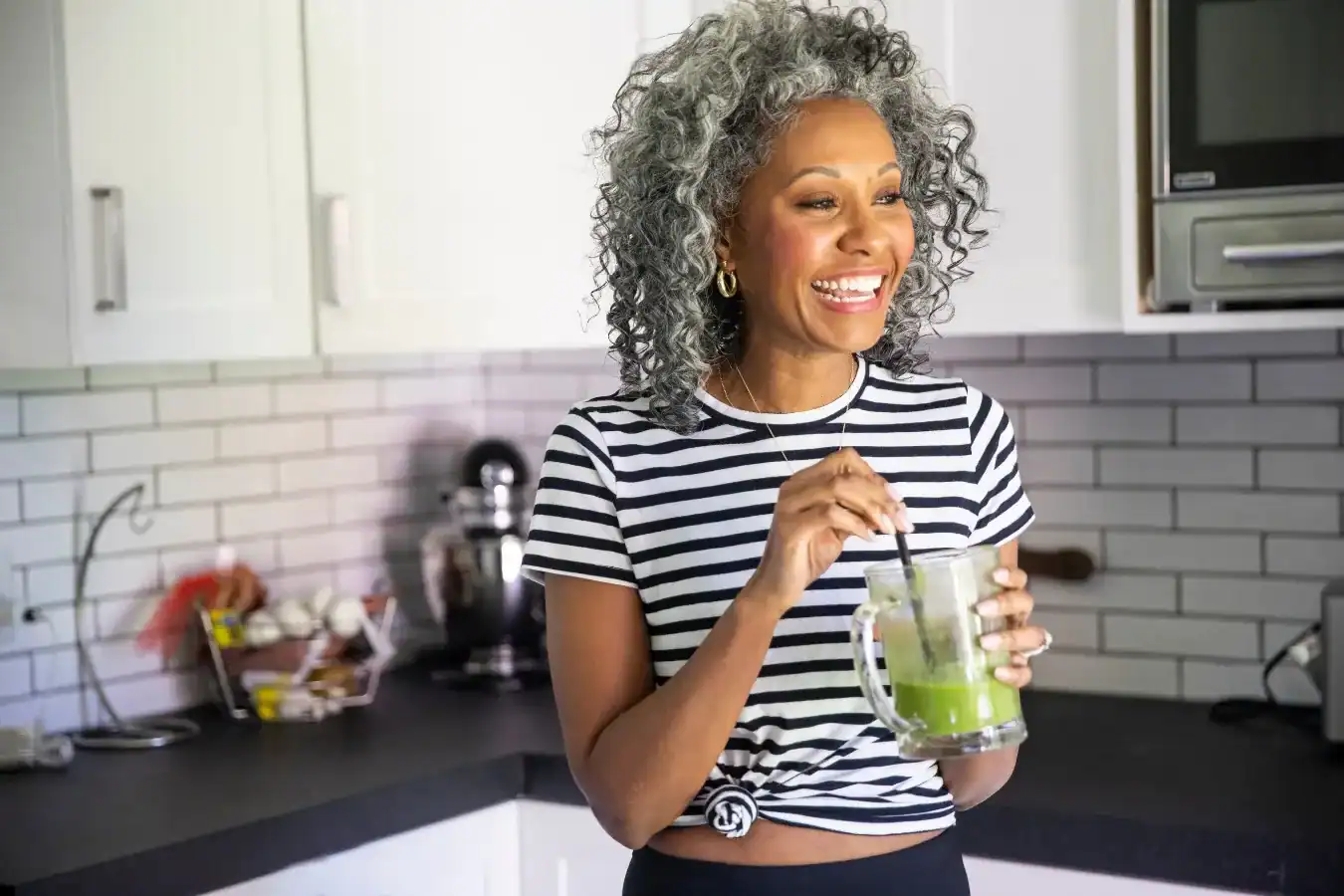 Weight Management Dietitians, In Good Nutrition, Smiling woman with curly gray hair in a striped shirt stands in a kitchen, holding and stirring a glass of green juice