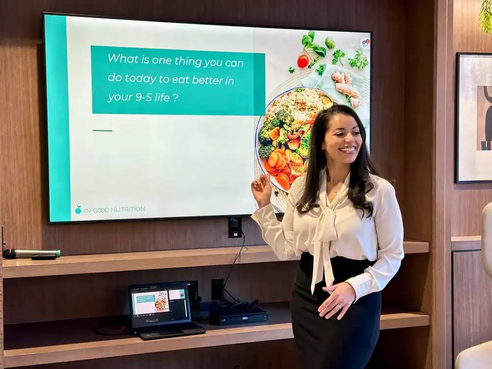 Registered Dietitian in business attire smiles and points to a screen displaying, “What is one thing you can do today to eat better in your 9-5 life?” with a photo of a healthy meal beside the text.